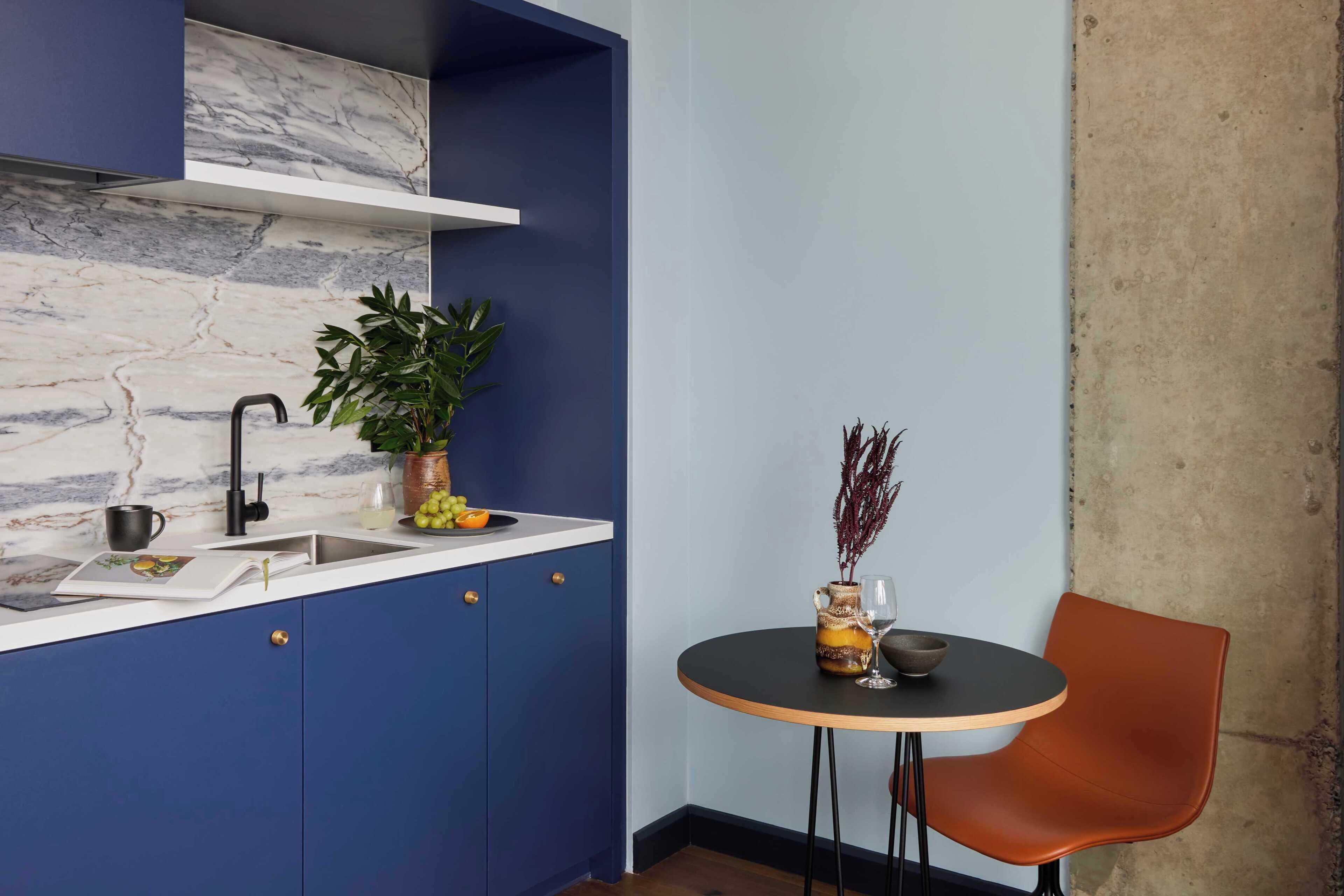 Une cuisine avec des meubles de couleur bleue, un pare-feu en marbre, un évier, une plante en pot, un vase contenant des plantes séchées et une table ronde avec une chaise.