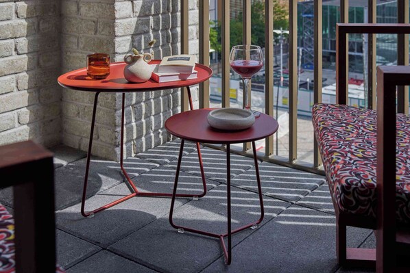 A balcony with a red table, a white teapot, a book, a glass of red wine, and a patterned sofa.