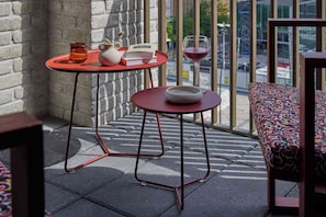 A balcony with a red table, a white teapot, a book, a glass of red wine, and a patterned sofa.
