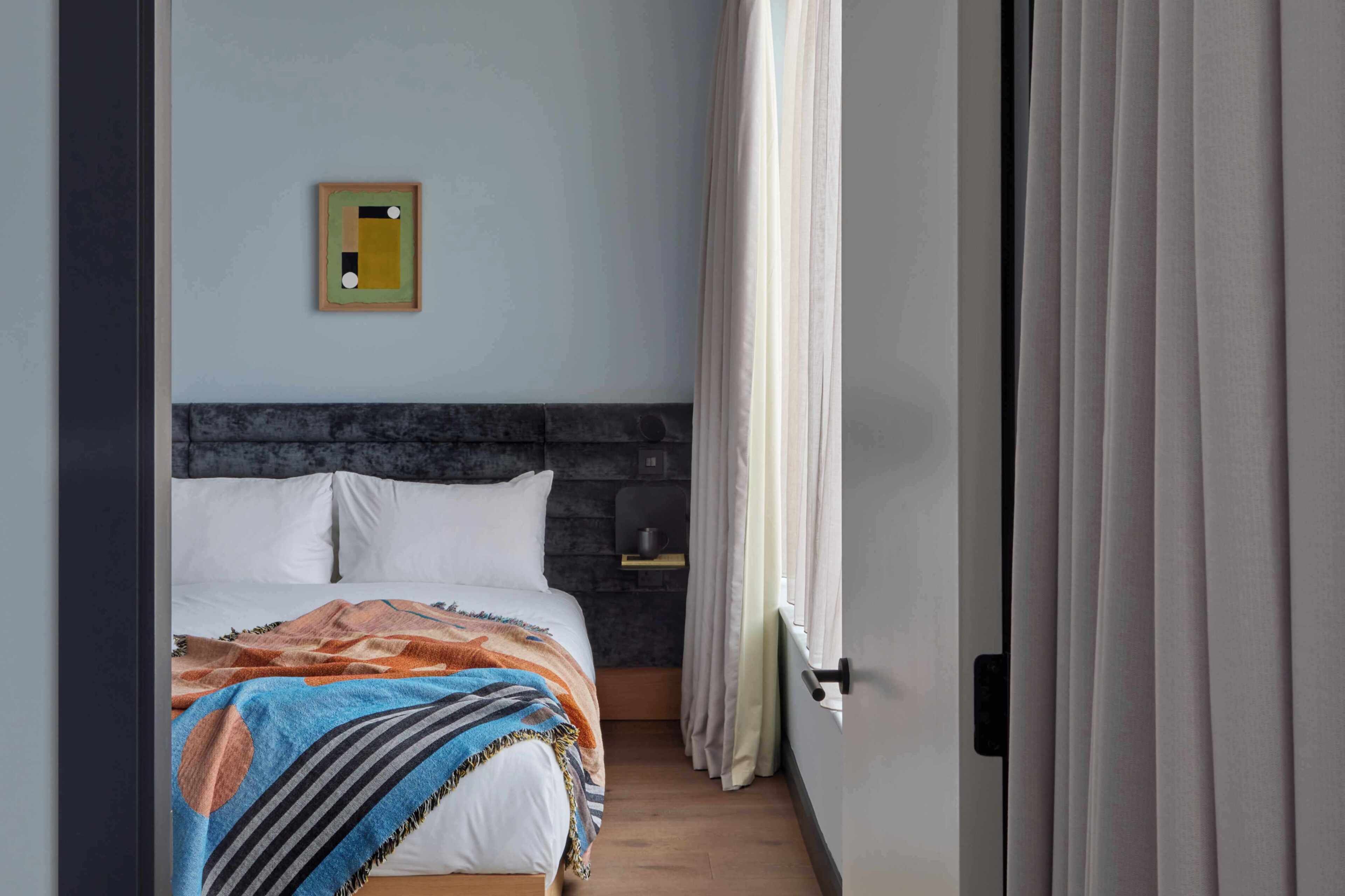 A bedroom with a bed, a headboard, a small shelf, and a window with curtains.