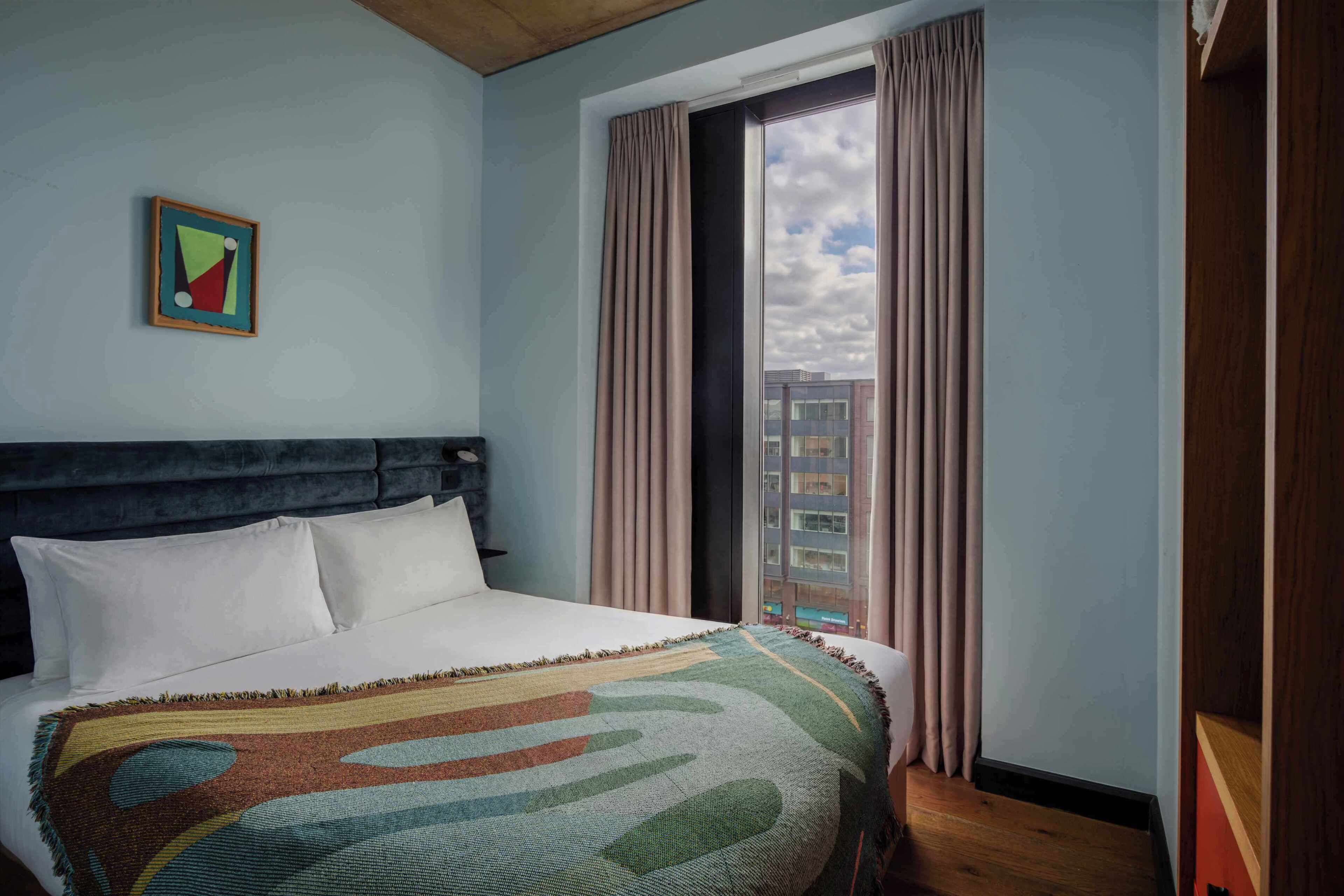 A hotel room with a bed, a window with curtains, a city view, and a framed picture on the wall.