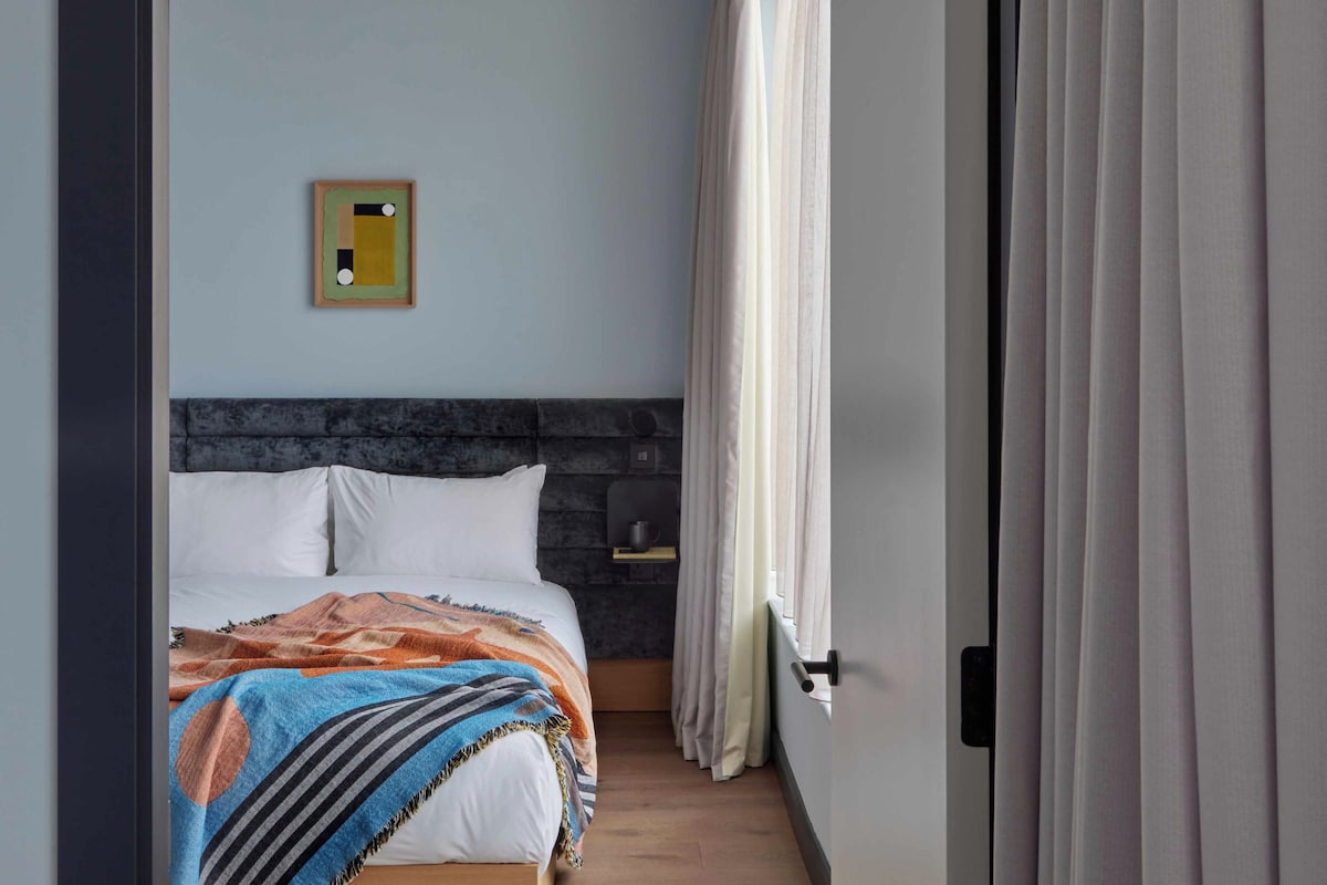 Un dormitorio con una cama, cabecera, repisa pequeña y ventana con cortinas.