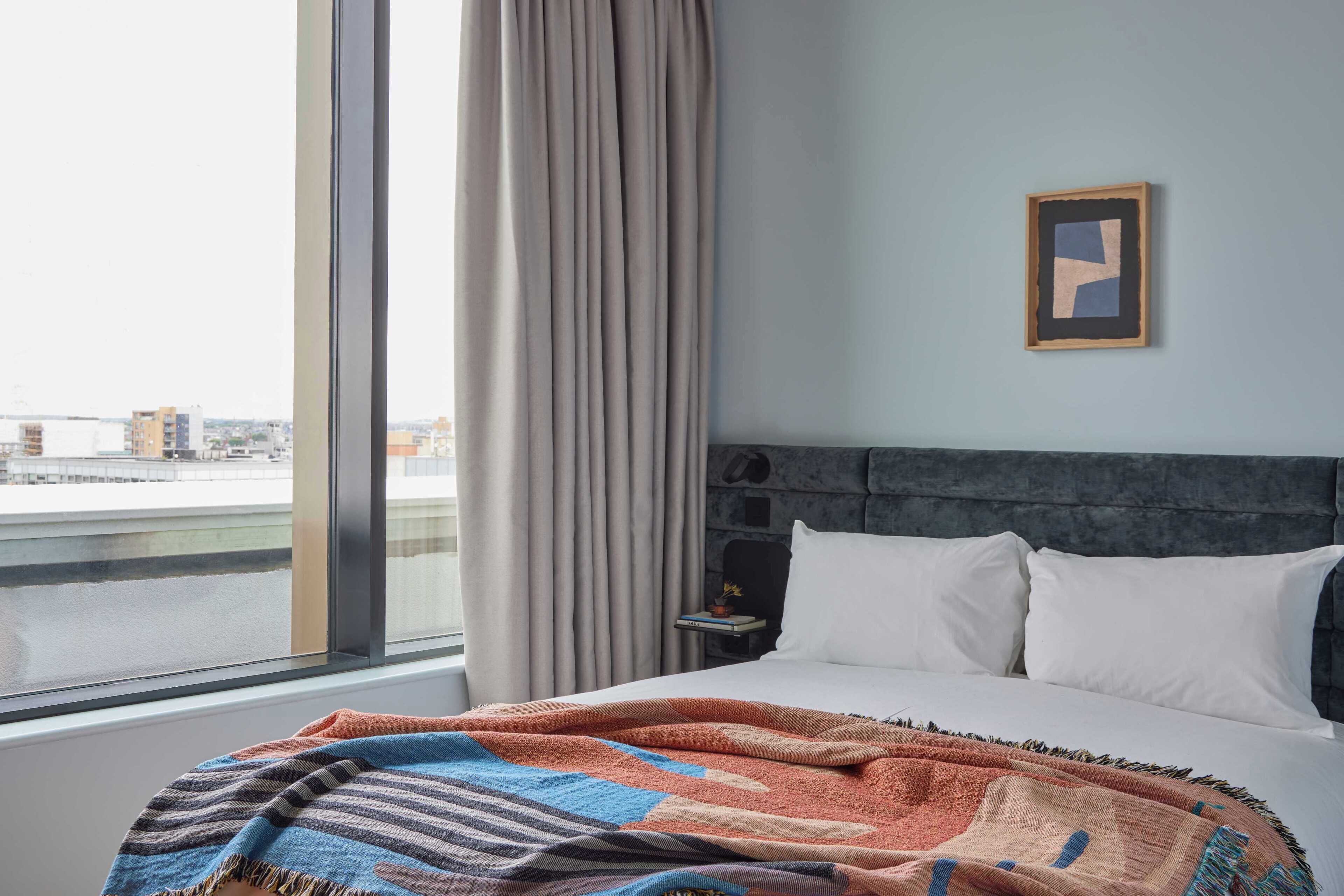 A hotel room with a large window, a bed with white linens, a patterned throw blanket, a framed picture on the wall, and a nightstand with a lamp.