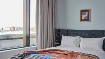 A hotel room with a large window, a bed with white linens, a patterned throw blanket, a framed picture on the wall, and a nightstand with a lamp.