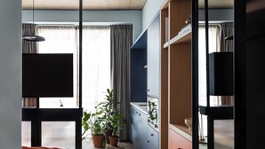 Una habitación moderna con cama, escritorio y un amplio ventanal con cortinas. Se observan plantas en macetas y un armario.