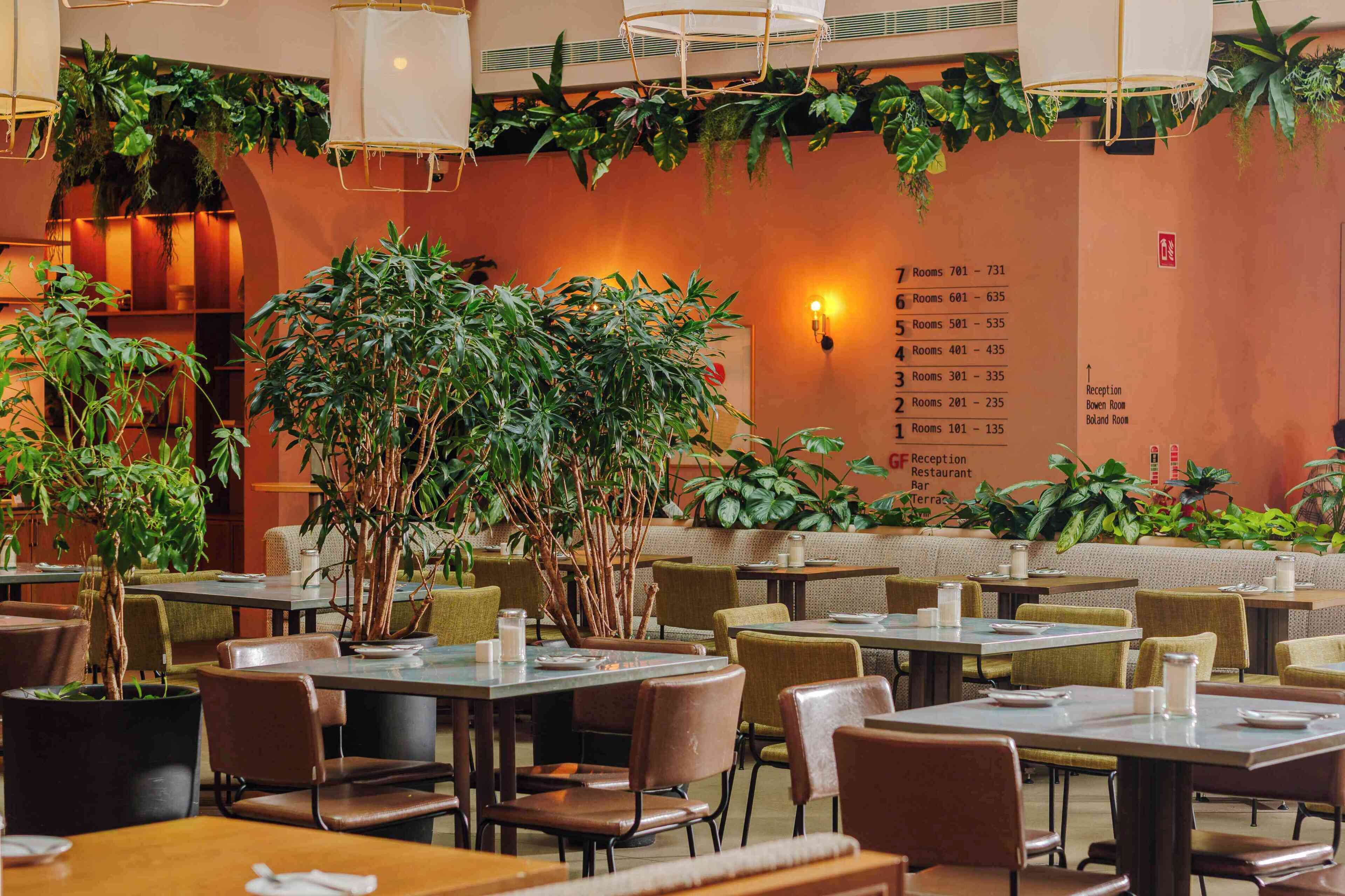 Un restaurant avec des tables et des chaises, un mur sur lequel figure une liste des numéros de chambre, et des plantes suspendues.