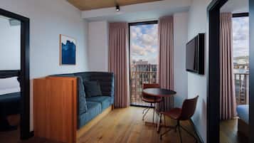 Ein modernes Hotelzimmer mit einer Sitzecke, einem kleinen Tisch, einem Sessel, einem Fernseher und einem großen Fenster mit Vorhängen.