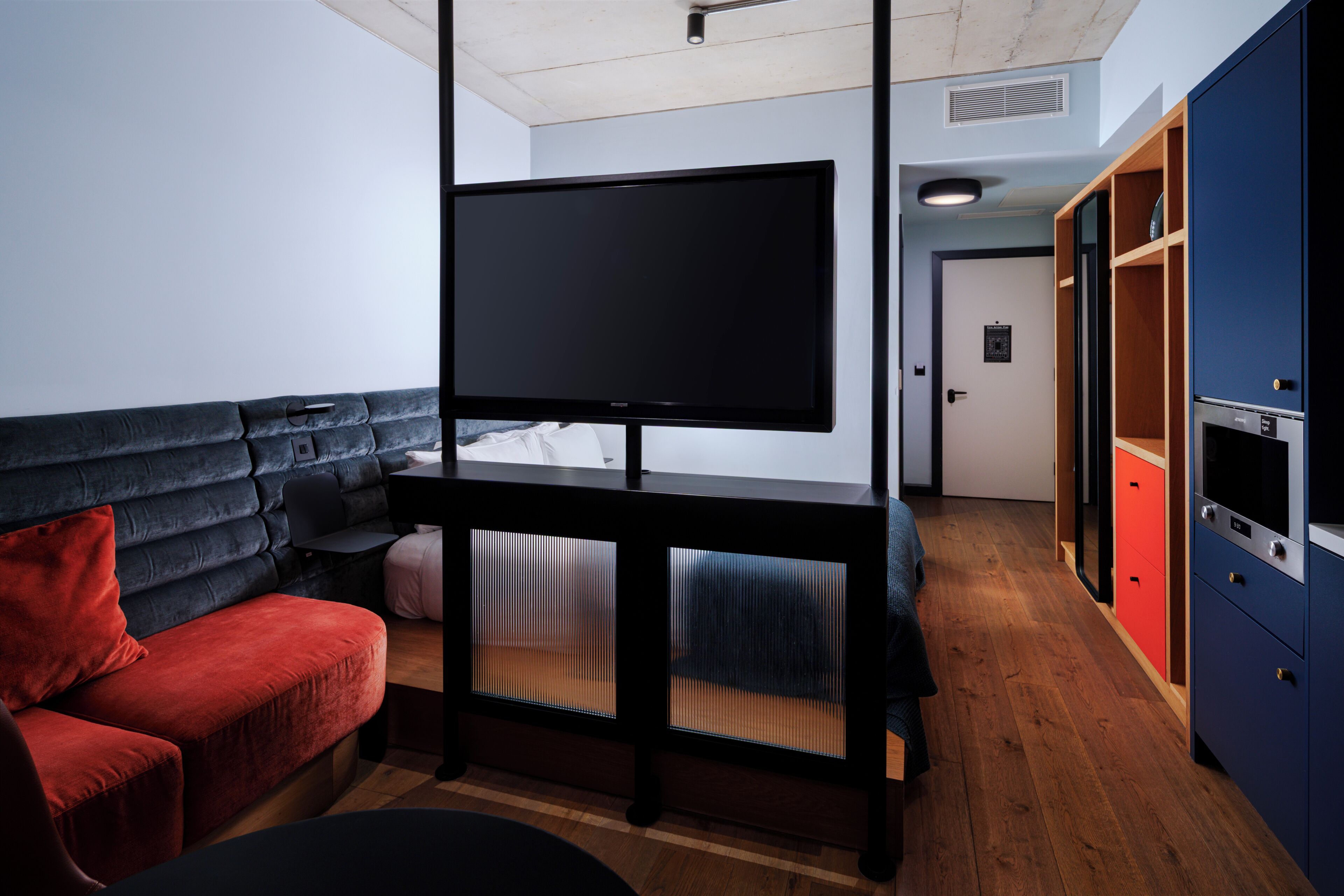 Quarto com televisão de tela plana em um suporte, sofá vermelho, piso de madeira e guarda-roupa com portas vermelhas e azuis.