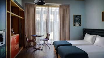 Habitación de hotel con cama, dos mesitas de noche, una silla, una mesita pequeña, un armario y una ventana con cortinas.
