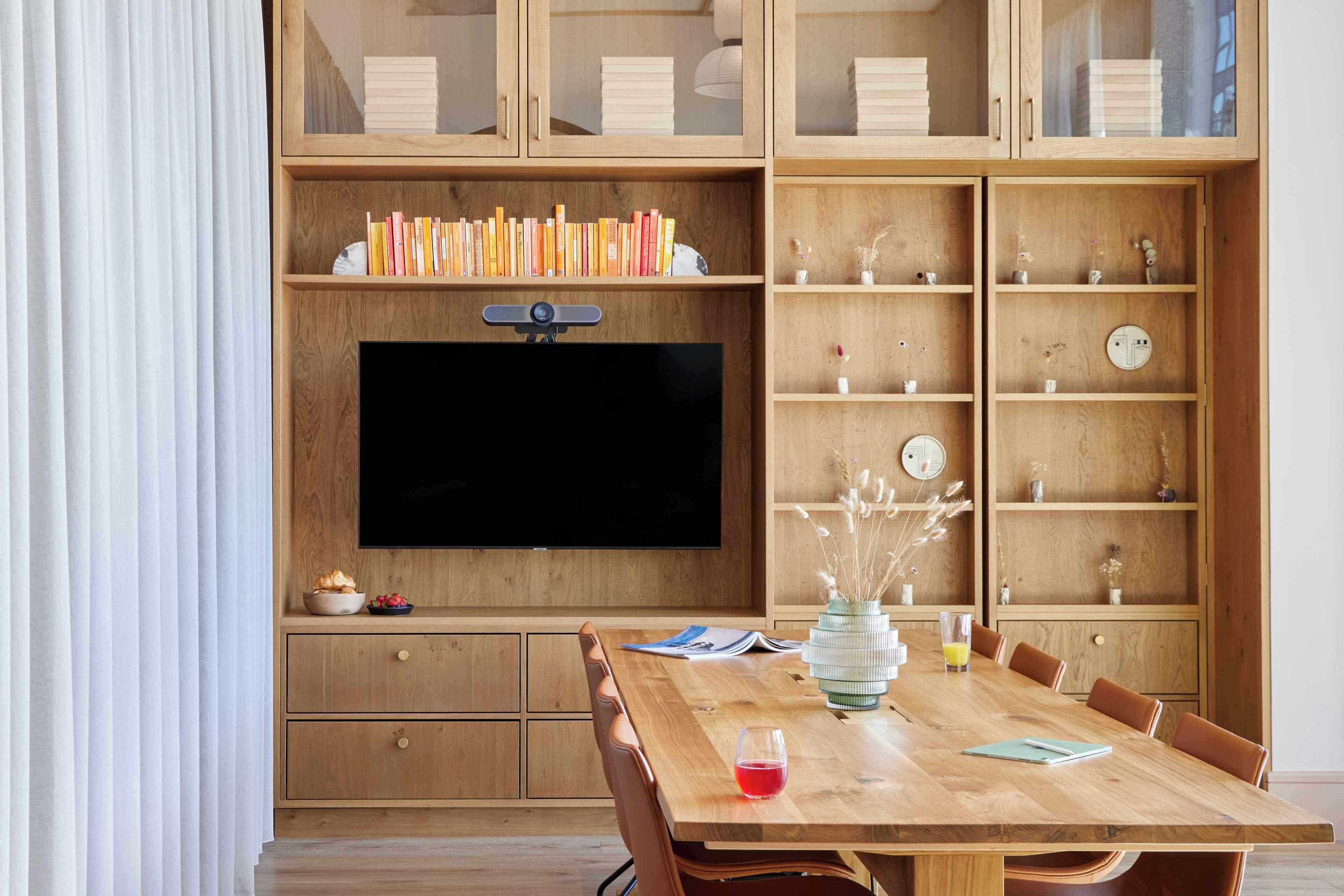 A dining area with a wooden table, chairs, a television mounted on a wooden cabinet, and shelves with books and decorative items.