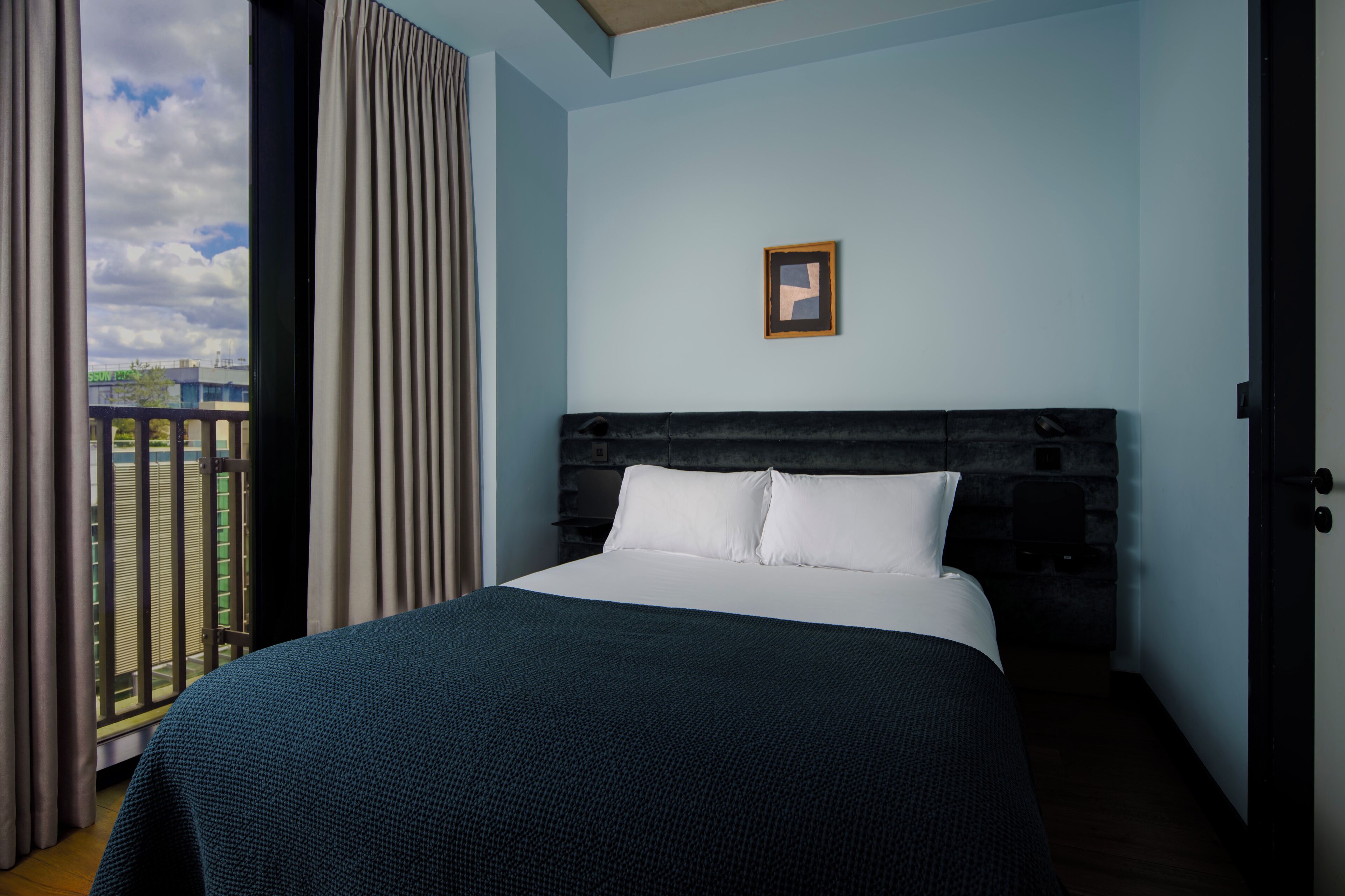 A hotel room with a bed, a framed picture on the wall, and a view of the outside.