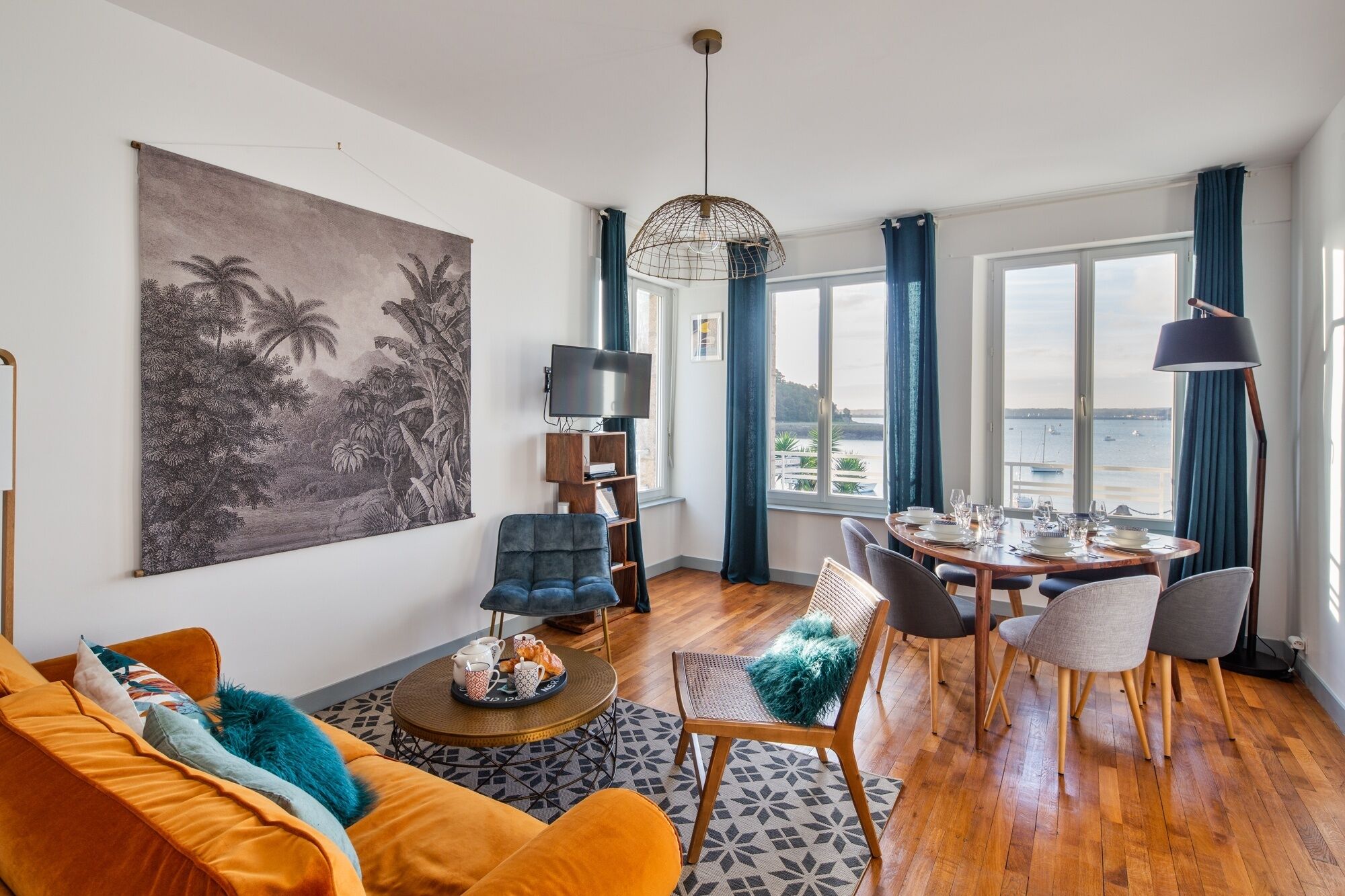 A living room with a large painting, a sofa, a coffee table, and a dining area with a view of the sea.