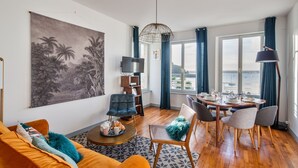 A living room with a large painting, a sofa, a coffee table, and a dining area with a view of the sea. - Le Zénith - avec vue mer (Saint-Malo)