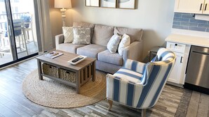 A living room with a sofa, armchair, coffee table, and a lamp. There are framed pictures on the wall, a kitchen area with cabinets and a refrigerator, and a view through a sliding glass door. - KING BED Oceanview 1BR Top Floor Villa (Hilton Head Island)