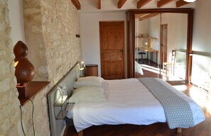 Une chambre avec un grand lit, une tête de lit en bois, un mur en pierre et une vue sur un balcon équipé d’un bureau et d’une chaise.