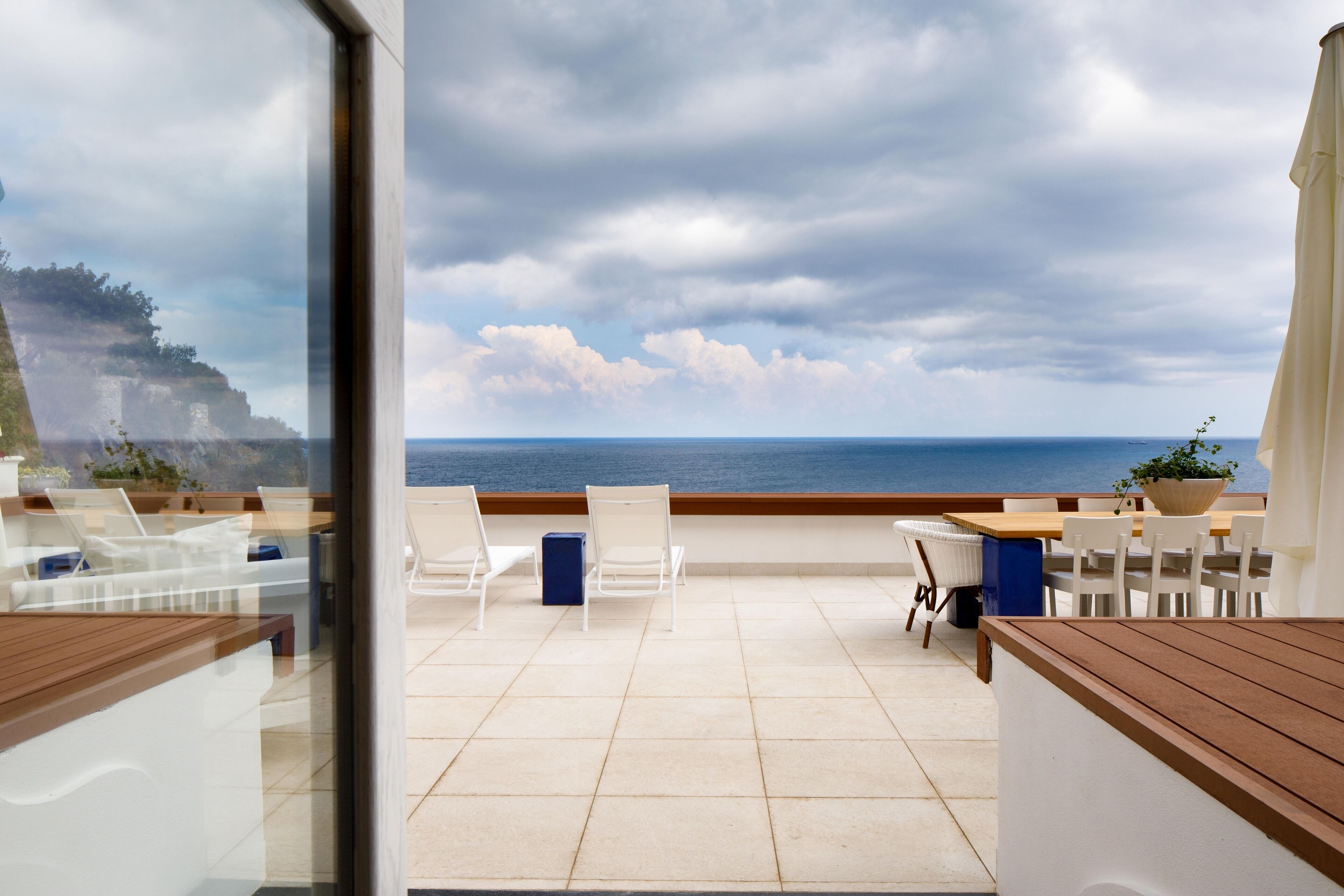 A terrace with a view of the sea, featuring a wooden table, white chairs, and a blue cabinet.