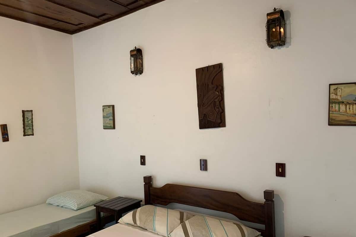 Una habitación con una cama grande, una cama más pequeña y un banco. Hay luces fijadas a la pared y un cuadro.