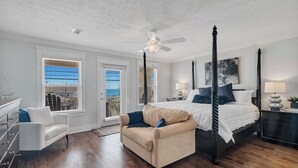 A four-poster bed with a ceiling fan, a beige sofa, and a white armchair in a room with wooden flooring and white walls. - Swell Life | Walk to Beach | Large Pool | Near 30A & Pier Park | Sleeps 12 (Panama City Beach)