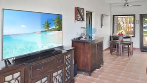A living room with a large flat-screen TV, wooden cabinets, a dining area with a table and chairs, and a view of the ocean through a window. - Golf View Rio Mar Villa (Río Grande)