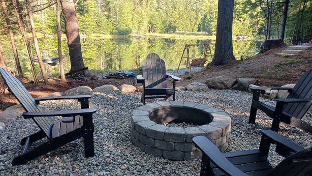 A fire pit surrounded by Adirondack chairs and a hammock in a forested area with a lake in the background. - Private lakeside cabin for those looking for peace and quiet in Brant Lake. (Brant Lake)