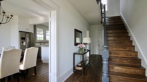 Amplia sala de estar con una escalera de madera, una mesa de comedor con sillas y una cocina al fondo.
