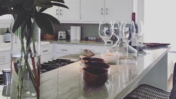 Una cocina moderna con armarios blancos, encimera de mármol, una planta grande, copas de vino y una botella de vino.