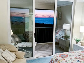 Une chambre à coucher comprenant un lit, une chaise, une table de chevet, une lampe et une vue sur l’océan, visible à travers une porte coulissante.