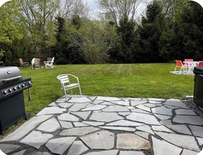 A stone patio with outdoor furniture, a grill, and a grassy yard with trees. - The Acadia Gateway House (Trenton)