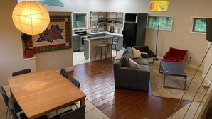 A modern kitchen with a central island, a dining area with a wooden table and chairs, a gray sofa with cushions, and a red chair. - The Acadia Gateway House (Trenton)