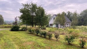 A house with a sloped roof, surrounded by a grassy area with small bushes and trees. - The Acadia Gateway House (Trenton)