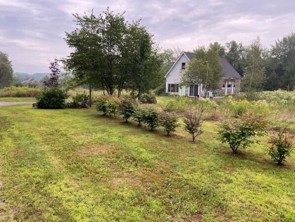 A house with a sloped roof, surrounded by a grassy area with small bushes and trees. - The Acadia Gateway House (Trenton)