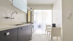 Eine moderne Küche mit weiß gefliestem Spritzschutz, dunklen Schränken, einer Edelstahlspüle und einem Gasherd. Im Hintergrund steht ein weißer Esstisch mit einem Stuhl.