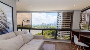 Ein modernes Wohnzimmer mit einer Couch, einem Schreibtisch und einem Sessel. Große Fenster bieten Blick auf die Stadtlandschaft.