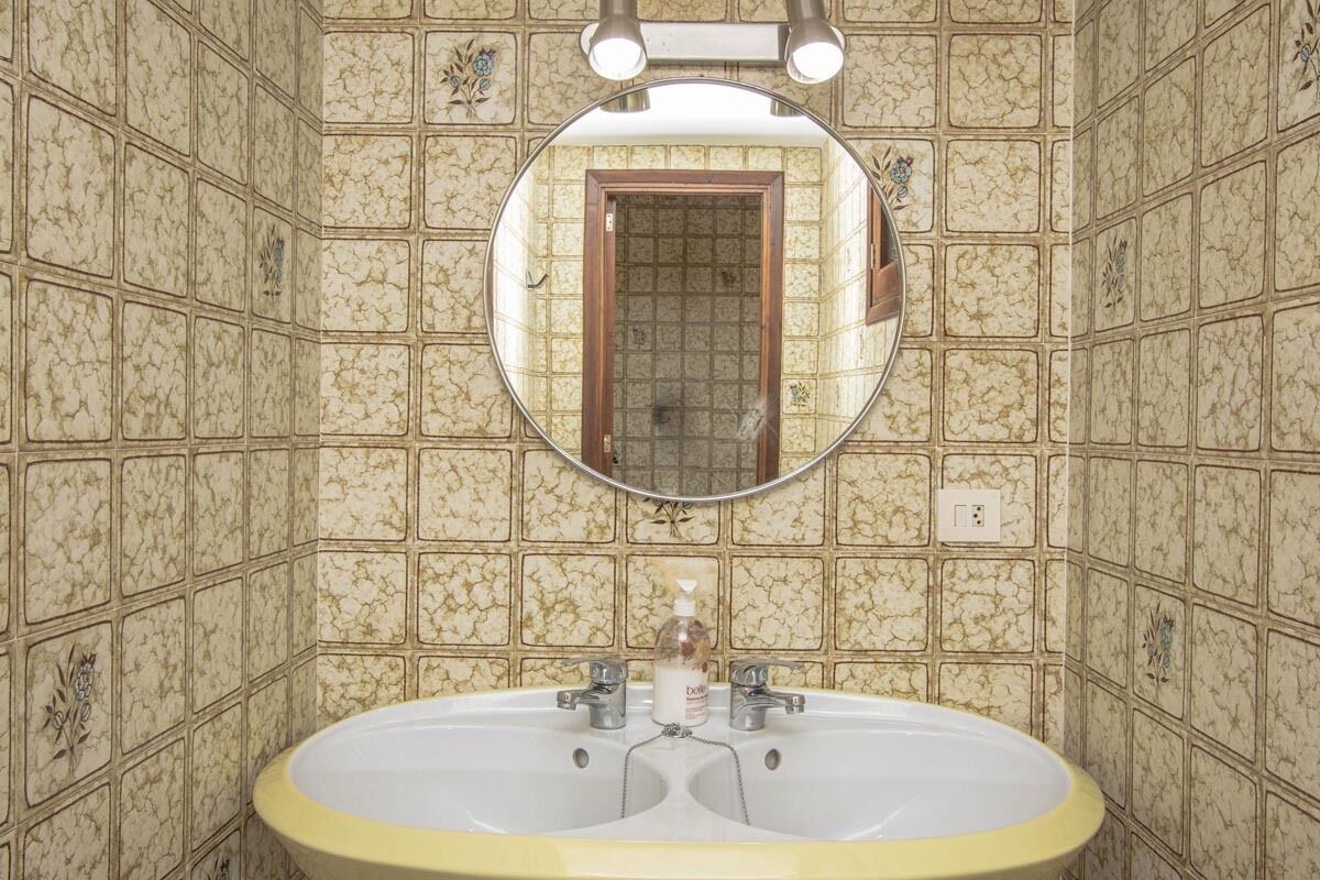 Un baño con un lavamanos ovalado grande, dos grifos y un espejo encima. Las paredes están cubiertas de azulejos con un diseño decorativo.