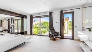 A modern hotel room with a large bed, a massage chair, a white vanity, and a balcony with a view of greenery. - Villa Cavall, Magnificent house 200 meters from the beach (Cala Sant Vicenç)