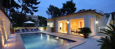 A swimming pool with lounge chairs, a covered patio area, and a staircase leading to a second floor.