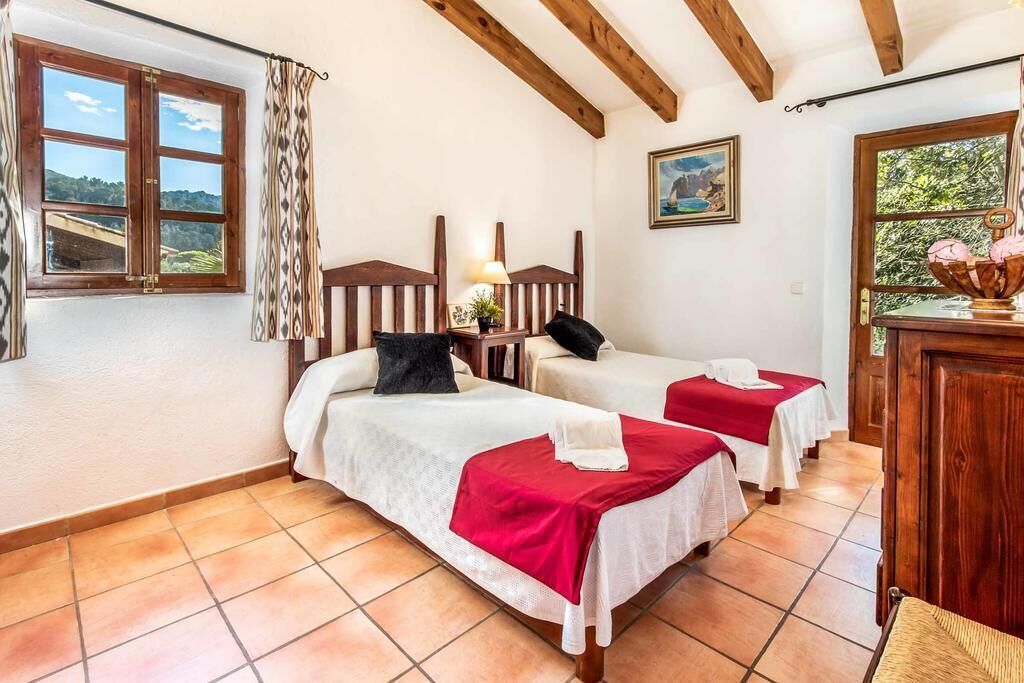A room with two beds, wooden beams on the ceiling, a window with curtains, and a painting on the wall.