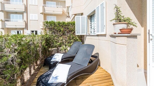 Een balkon met twee loungestoelen en een kleine tafel, omgeven door groen en een gebouw met meerdere ramen en balkons.