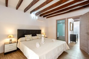 A bedroom with a large bed, wooden beams on the ceiling, a nightstand with a lamp, and a bathroom visible through an open door.