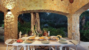 A stone-arched outdoor dining area with a table set for breakfast, surrounded by greenery and a large tree trunk. - Villa Alordes, Newly renovated house with mountain views (Pollença)