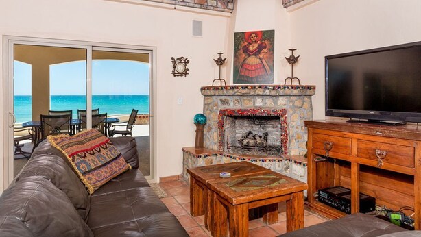 A living room with a stone fireplace, a television, and a view of the ocean. - Casa San Miguel Beachfront House (Puerto Peñasco)