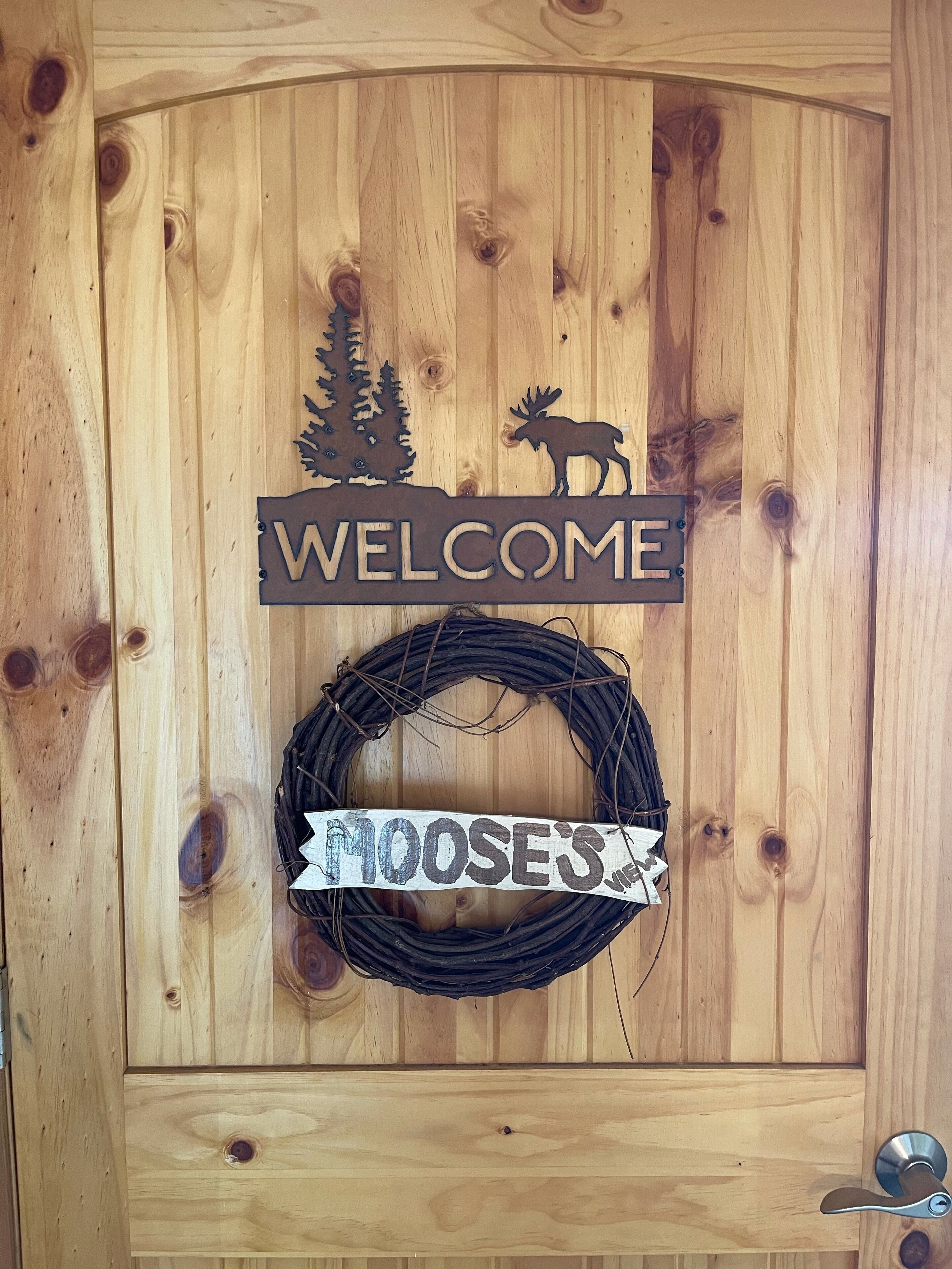 Een houten deur met een 'WELCOME' bord en een 'MOOSE'S' bord, een krans en een houten paneel met een metalen handvat.