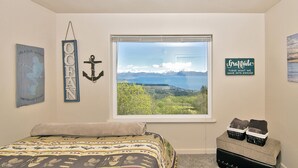Une chambre à coucher avec un lit, une fenêtre donnant sur un paysage de montagnes et d’eau, et des éléments de décoration murale comprenant un panneau et une ancre.