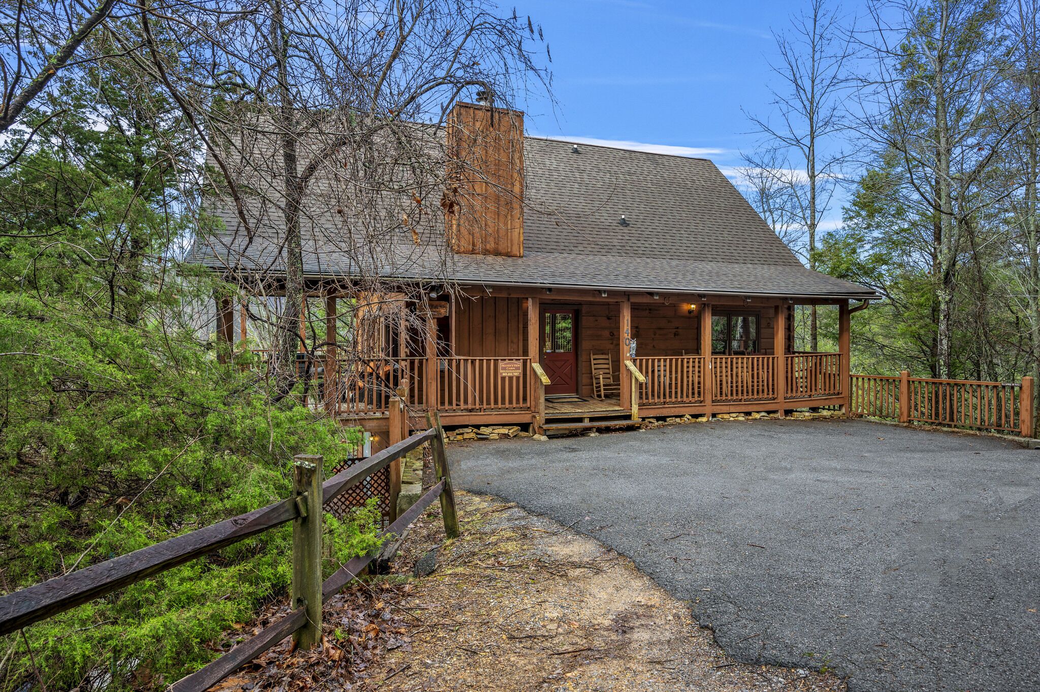 A rustic cabin with a porch, surrounded by trees and a gravel driveway.