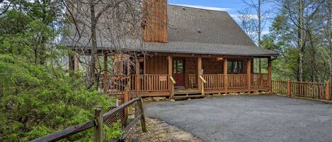 A rustic cabin with a porch, surrounded by trees and a gravel driveway.