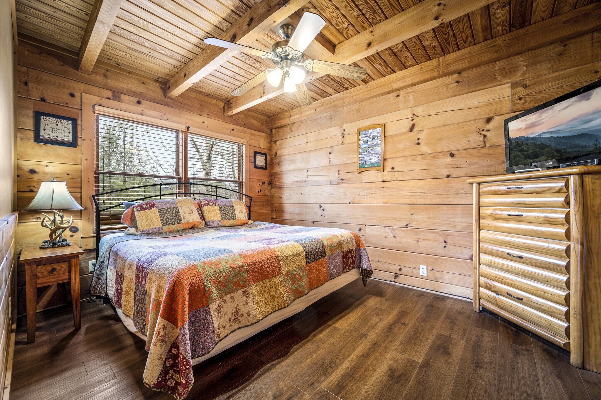 Un dormitorio de cabaña de madera con una cama, un ventilador de techo, un aparador y un televisor sobre un mueble de madera.