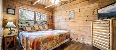 Un dormitorio de cabaña de madera con una cama, un ventilador de techo, un aparador y un televisor sobre un mueble de madera.