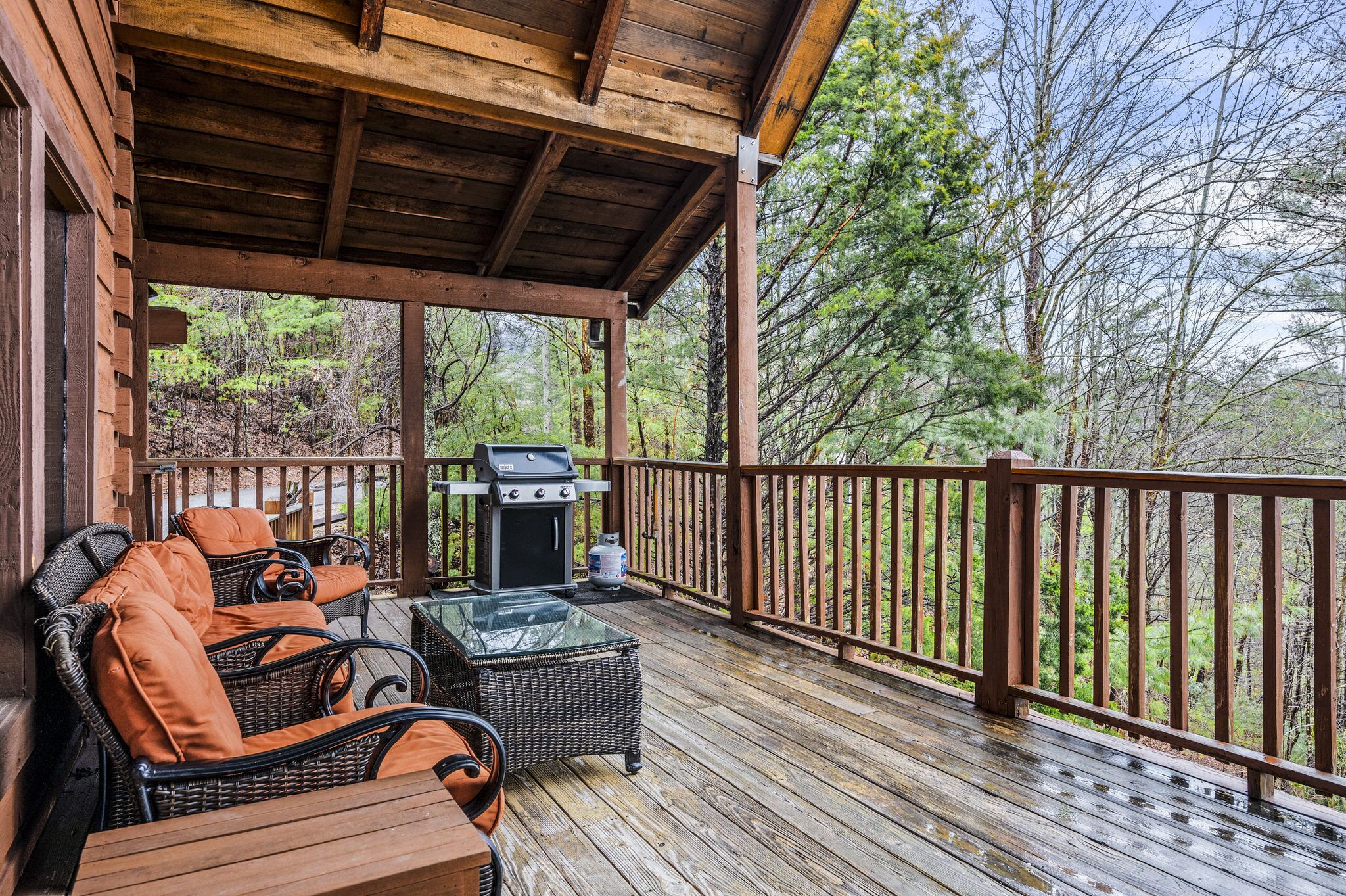 Une terrasse couverte, aménagée avec des meubles en osier et dotée d’un barbecue, qui offre une vue sur une zone boisée.