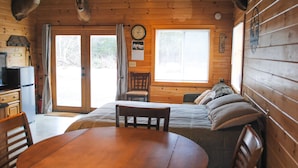 Ein Interieur einer Holzhütte mit einem Holztisch, Stühlen, einem Bett mit Kissen, einer Uhr und einem aufgehängten Hirschkopf.