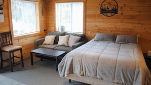 Ein gemütliches Zimmer mit einem großen Bett, einer Couch und einem Sessel. Zwei Fenster mit Jalousien, eine Holzwand mit einer Willkommensschild und ein kleiner Tisch sind ebenfalls vorhanden.